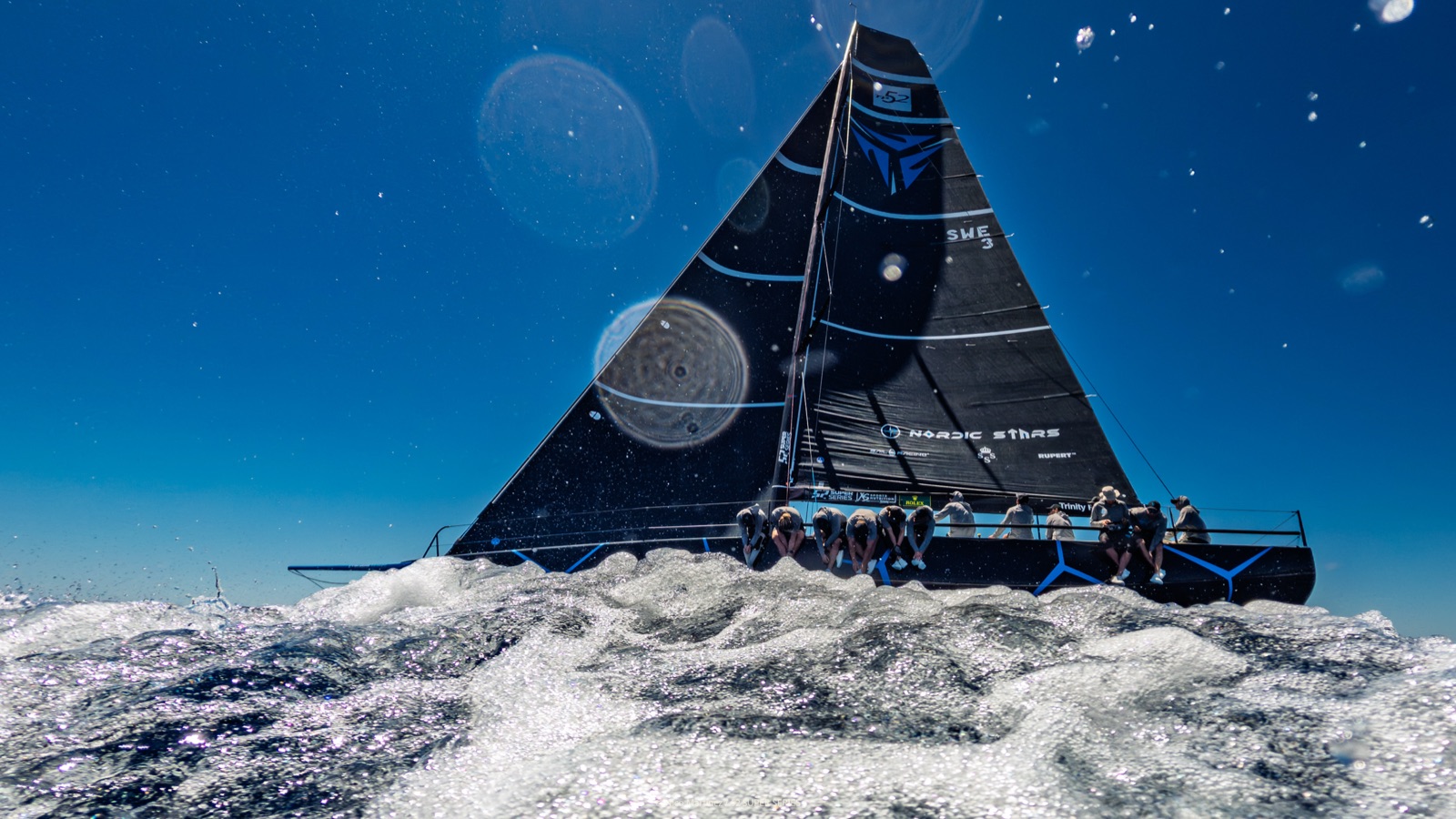 Water-level view of Nordic Stars under full sail, wake foaming past the camera